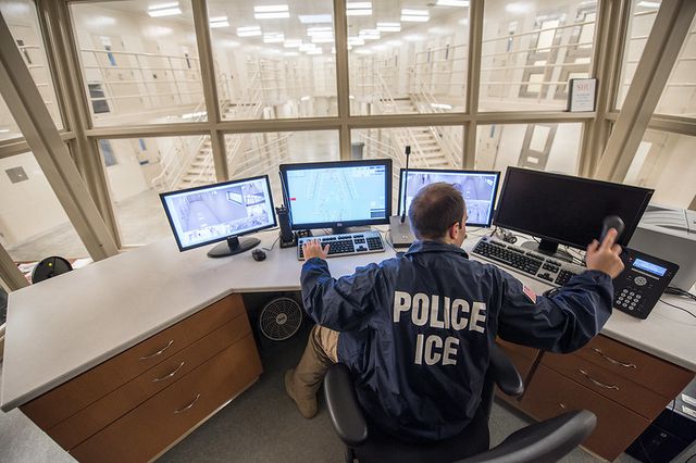 A security guard sits before four computer screens showing surveillance video of corridors inside an ICE detention facility in Batavia, NY.